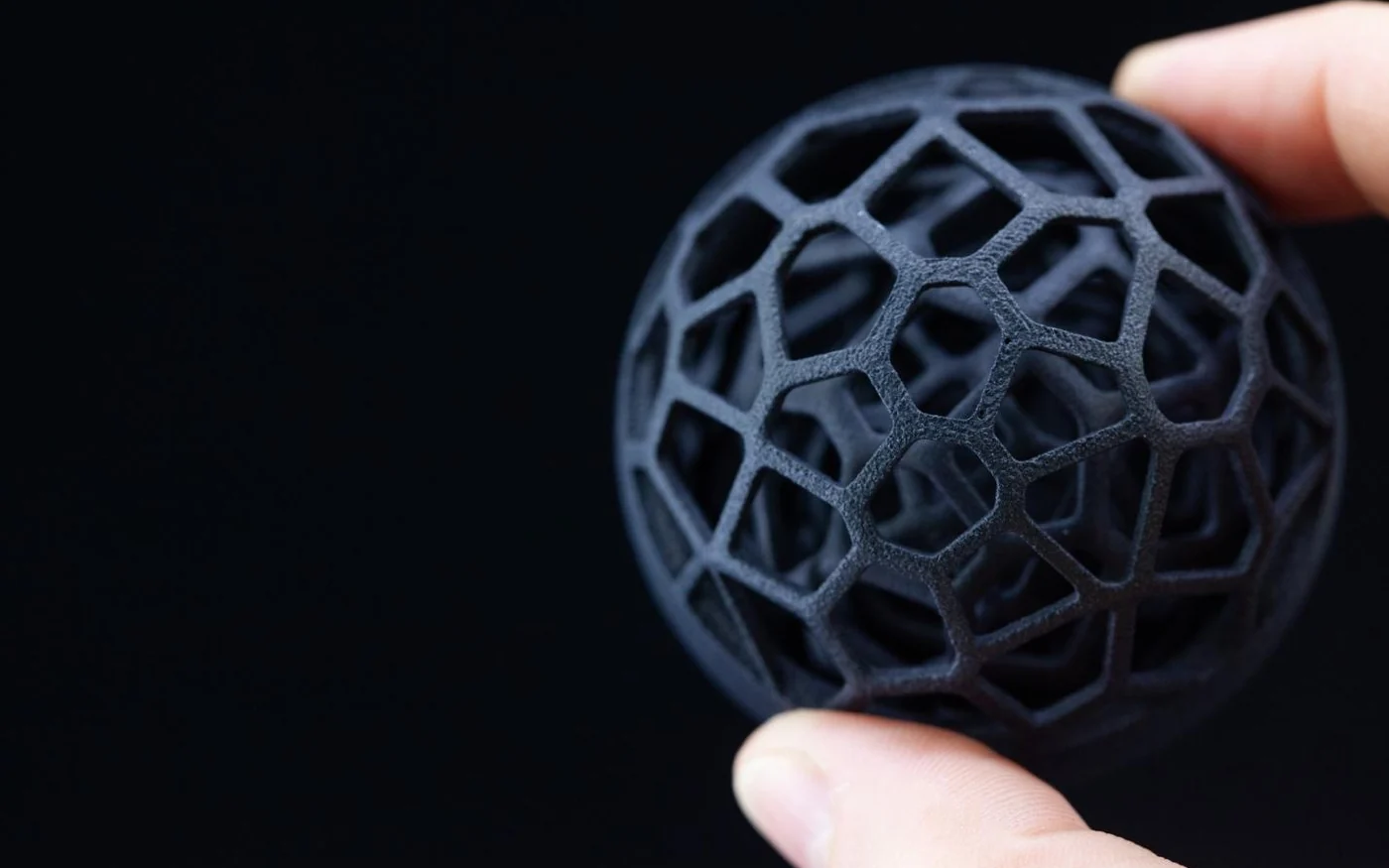 A 3D printed lattice sphere held between finger and thumb — geometry designed for additive manufacturing